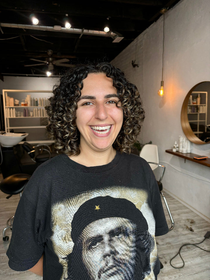 Dark curly hair with highlights on a person smiling at the camera in a hair salon.