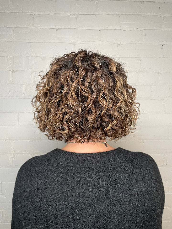 Highlighted curly hair bob from the back against a white wall.