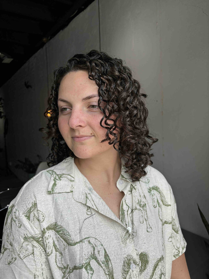 Woman with curly hair wearing a light-colored shirt with horse pattern indoors.